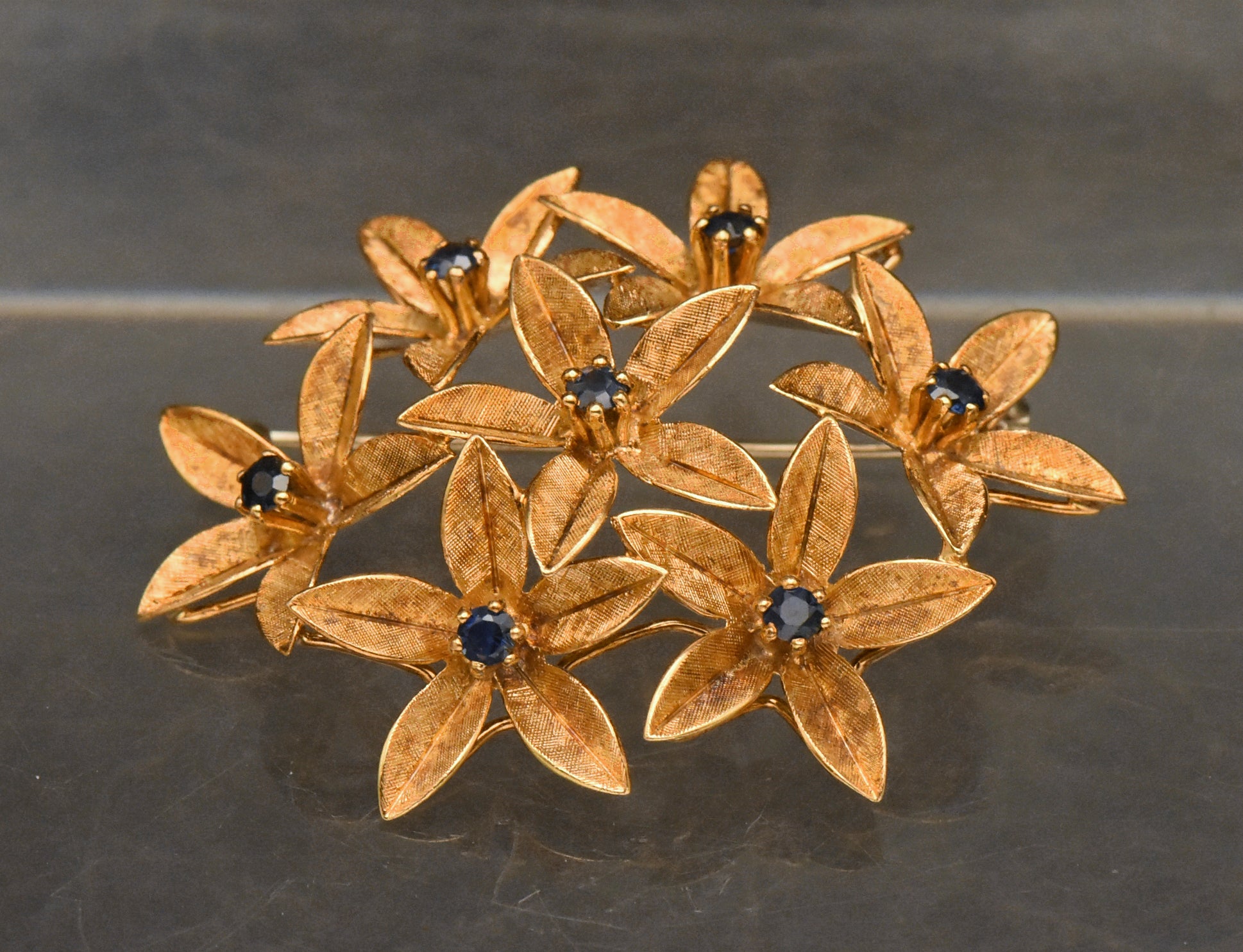 Stunning vintage antique handcrafted 18k gold brooch consisting of seven flowers with florentine finish, each set with a round cut sapphire.