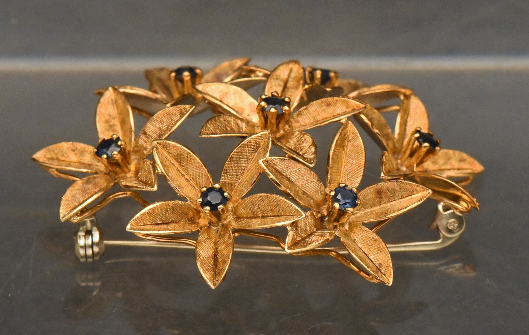 Stunning vintage antique handcrafted 18k gold brooch consisting of seven flowers with florentine finish, each set with a round cut sapphire.