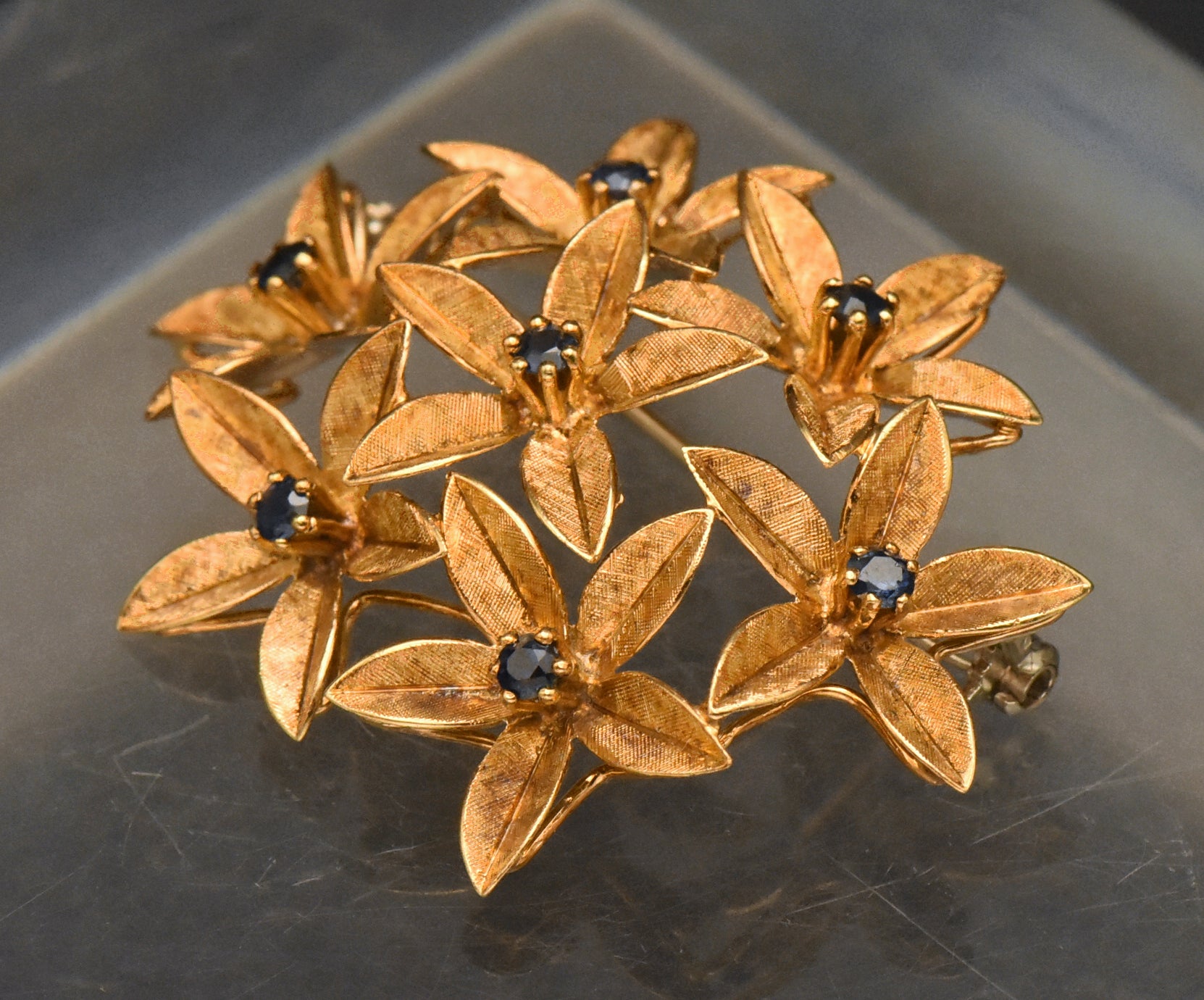 Stunning vintage antique handcrafted 18k gold brooch consisting of seven flowers with florentine finish, each set with a round cut sapphire.